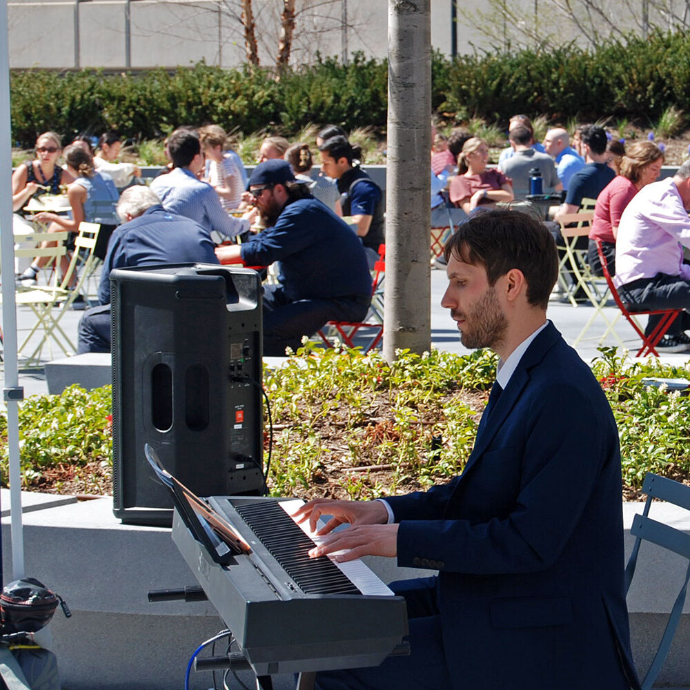 a man plays piano in a park while officer workers enjoy lunch on the patio listening to live music